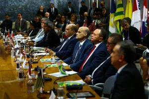 Autoridades brasileiras participam de reunião oficial com bandeiras estaduais e do Brasil ao fundo.
