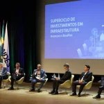 Painel de especialistas debate superciclo de investimentos em infraestrutura no Brasil durante evento com palco e telão ao fundo.