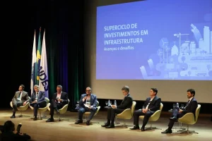 Painel de especialistas debate superciclo de investimentos em infraestrutura no Brasil durante evento com palco e telão ao fundo.