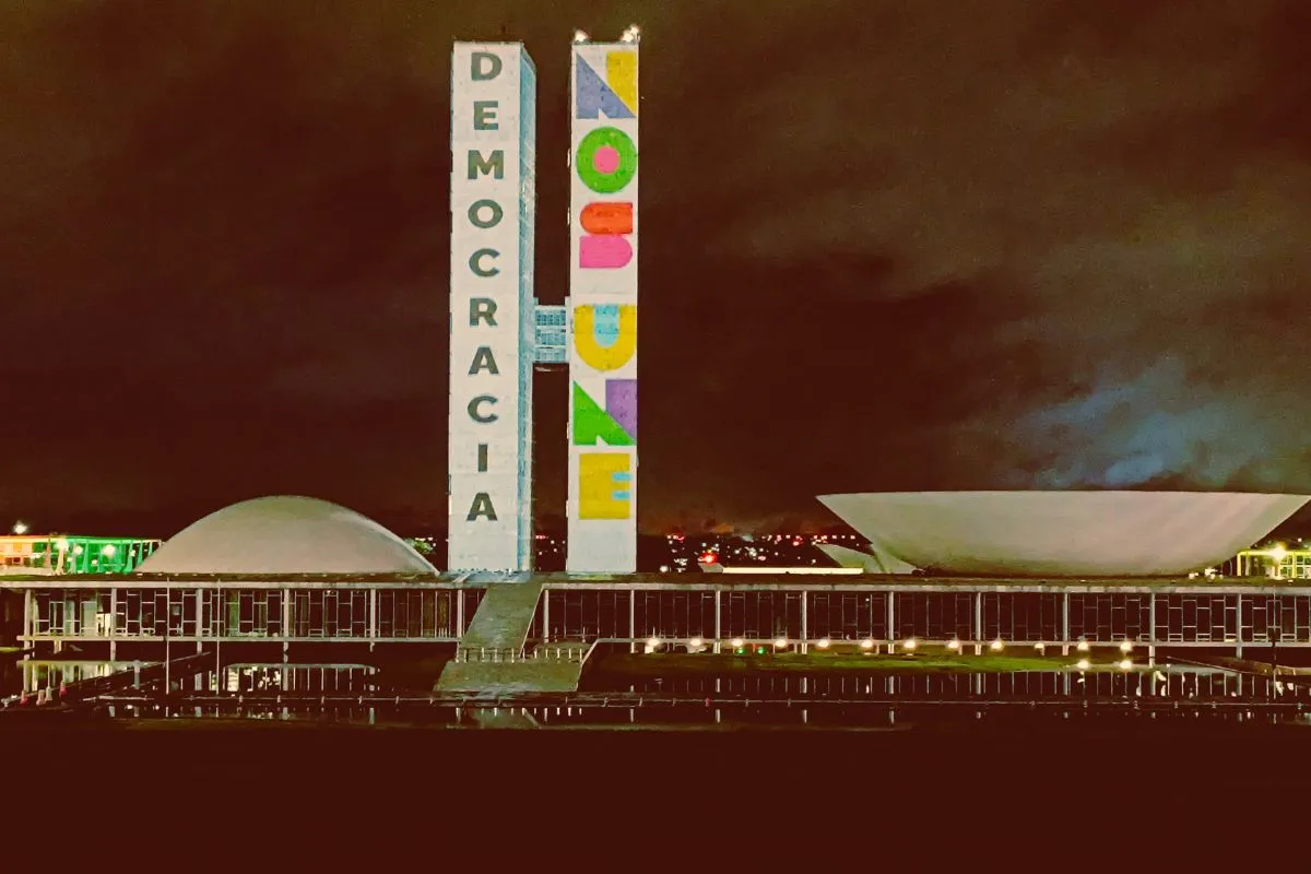 Congresso Nacional em Brasília iluminado à noite com projeções das palavras Democracia e No More nas torres.