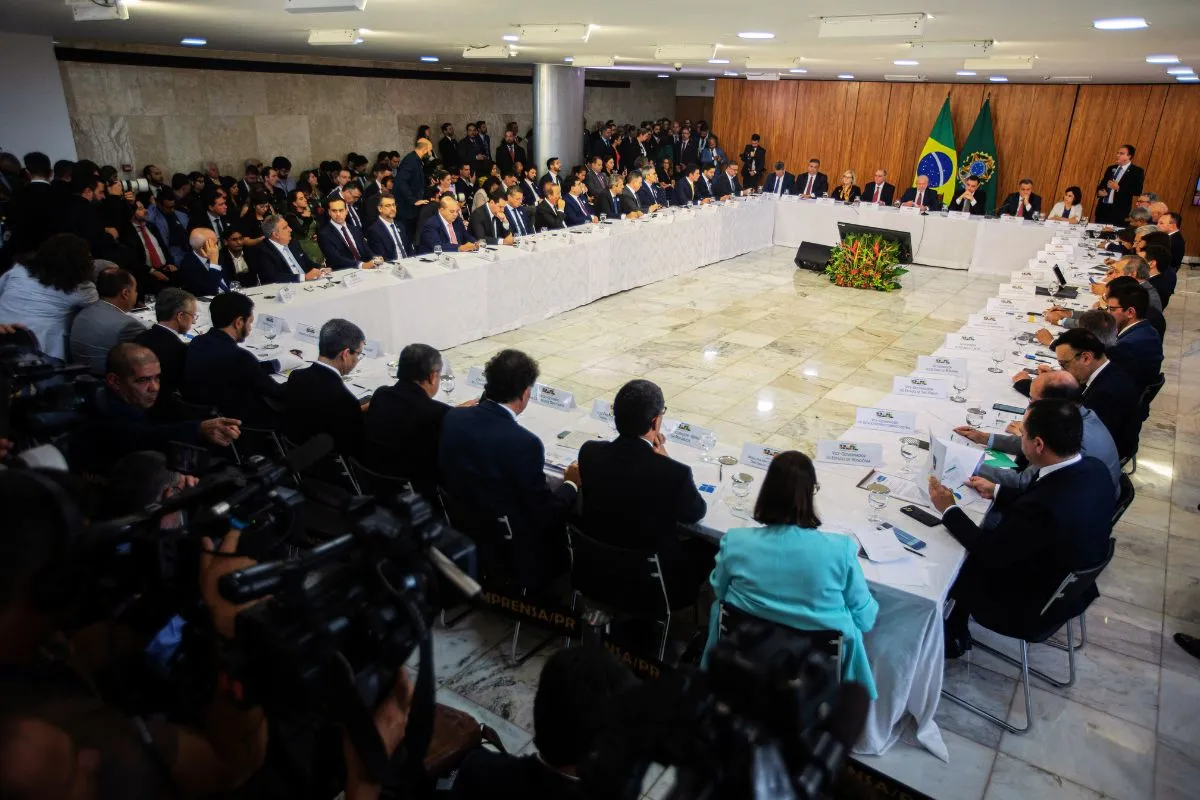 Autoridades do Governo Federal participam de reunião ministerial em mesa ampla no Palácio do Planalto, com bandeiras do Brasil ao fundo.