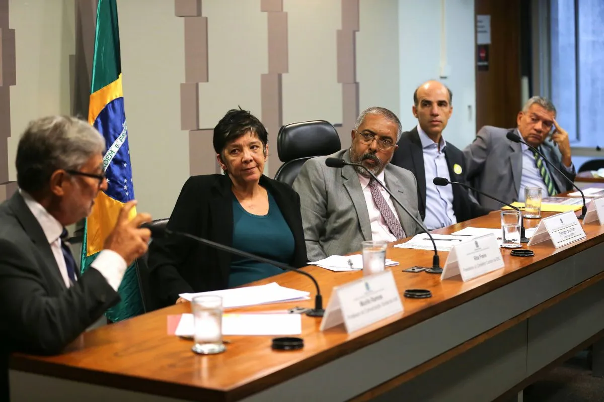 Autoridades sentadas em mesa de reunião com bandeira do Brasil ao fundo durante debate institucional.