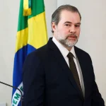 Autoridade brasileira de terno em pronunciamento oficial com bandeira do Brasil ao fundo.