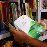 Pessoa segura livro infantil ilustrado em frente a estante de biblioteca escolar.