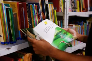 Pessoa segura livro infantil ilustrado em frente a estante de biblioteca escolar.