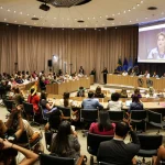 Participantes acompanham audiência pública sobre políticas para mulheres e saúde em auditório com transmissão em telão.