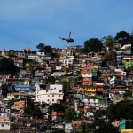 Helicóptero sobrevoa comunidade em área de favela no Brasil com casas densamente construídas em morro.