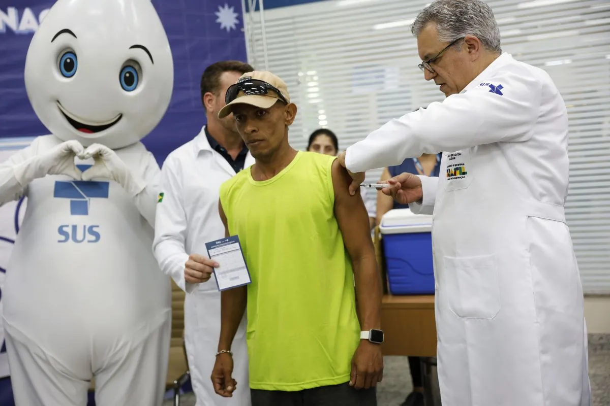 Profissional de saúde aplica vacina em cidadão durante campanha do SUS com presença de mascote institucional.