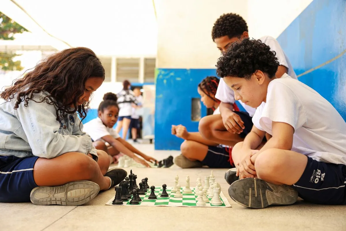 Crianças jogam xadrez no pátio de escola pública durante atividade educativa.