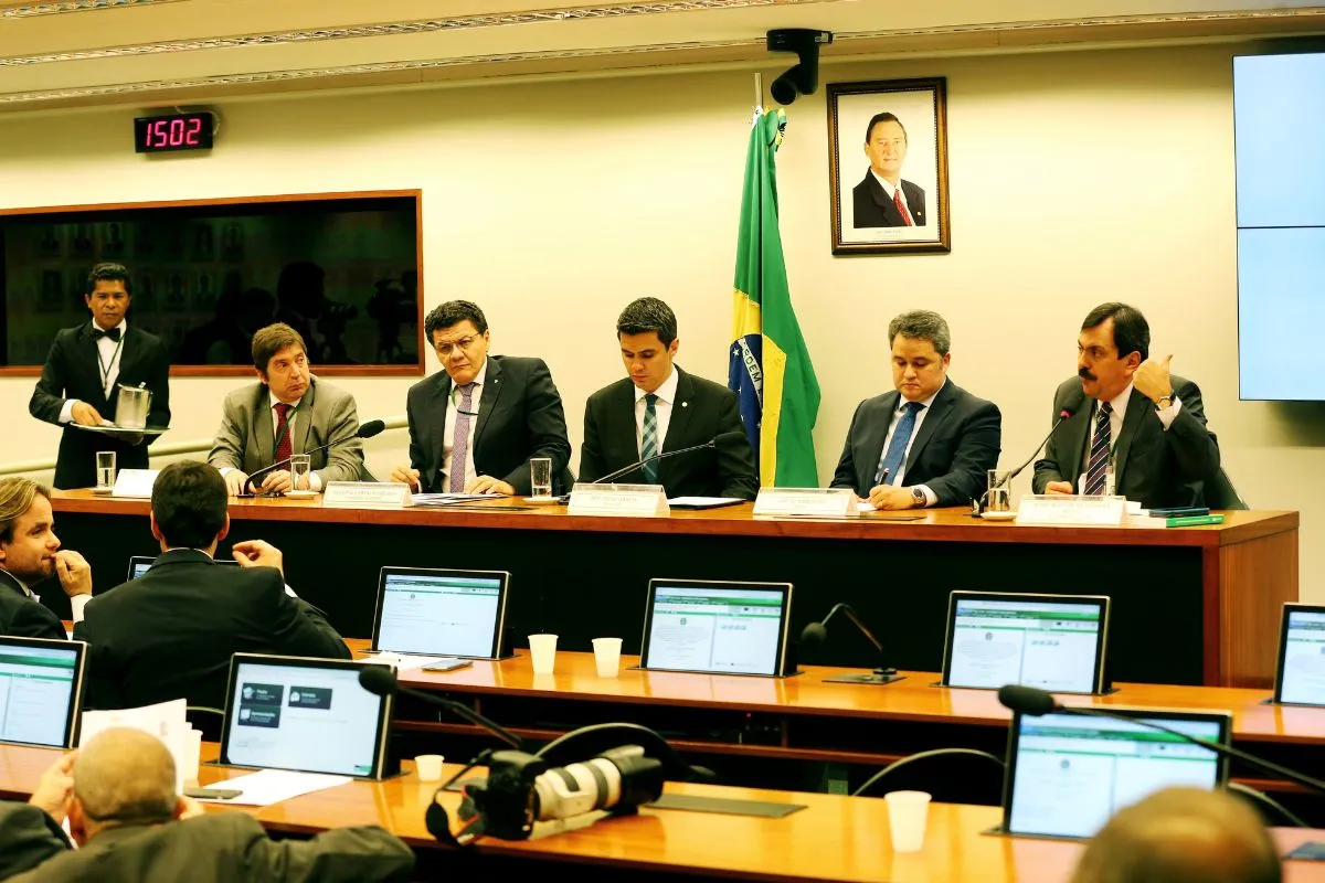 Autoridades reunidas em comissão parlamentar durante sessão institucional com computadores e documentos