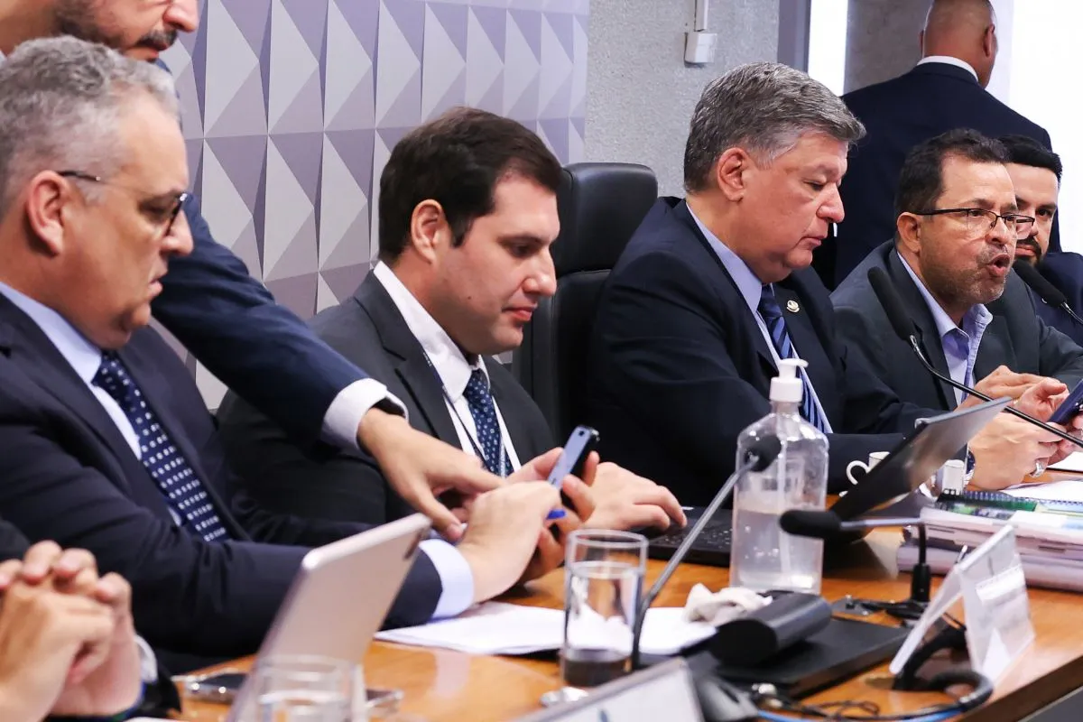 Autoridades sentadas em mesa de comissão durante inquérito institucional, utilizando celulares e documentos
