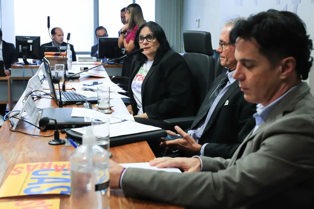 Audiência em comissão parlamentar com participantes sentados à mesa, uso de microfones e documentos durante debate institucional.