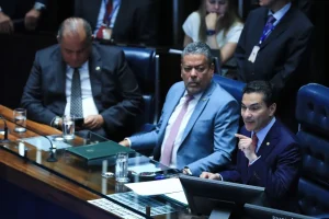 Senadores participam de sessão no plenário do Senado Federal, com parlamentares sentados à mesa e um deles discursando.