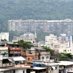 Comunidade com casas simples em primeiro plano e prédios residenciais ao fundo em área urbana do Brasil.