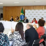 Autoridades reunidas em mesa oficial com bandeira do Brasil durante reunião ministerial.