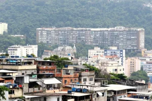 Comunidade com casas simples em primeiro plano e prédios residenciais ao fundo em área urbana do Brasil.