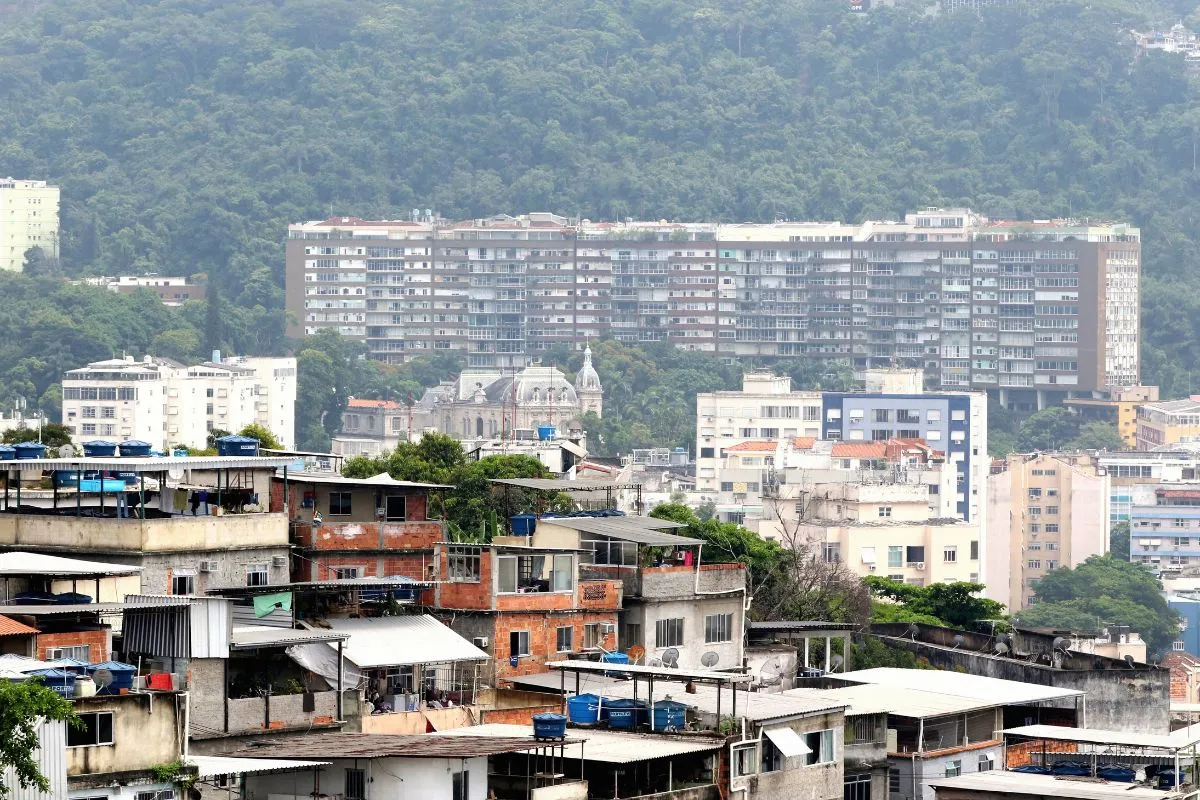 Comunidade com casas simples em primeiro plano e prédios residenciais ao fundo em área urbana do Brasil.