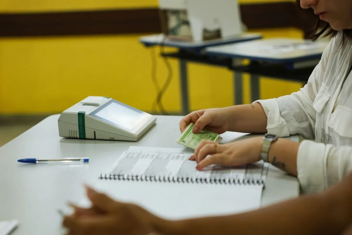 Mulher entregando documento de identificação para mesária em seção eleitoral com urna eletrônica ao lado