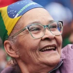 Senhora idosa sorrindo com óculos e usando bandana com a bandeira do Brasil durante manifestação patriótica