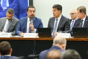 Autoridades políticas participam de audiência pública na Câmara dos Deputados, com parlamentares sentados à mesa e um deles falando ao microfone.