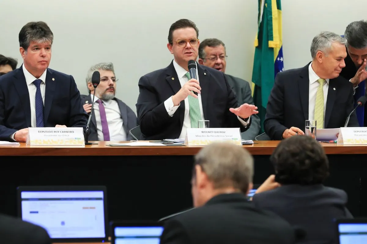 Autoridades sentadas à mesa durante audiência pública, com microfone e bandeira do Brasil ao fundo.