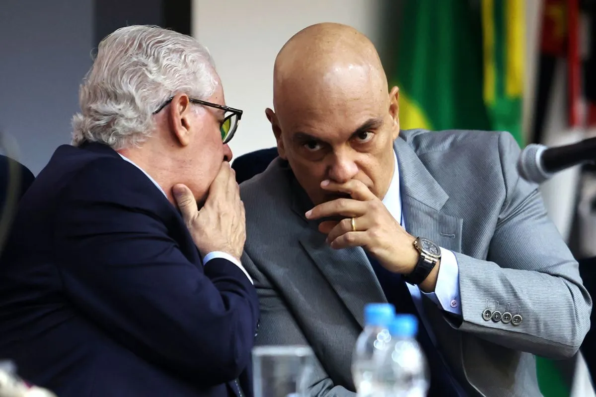Duas autoridades conversando de forma reservada durante reunião institucional, sentadas à mesa em ambiente oficial.
