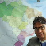 Homem de terno e óculos em frente a mapa do Brasil, representando dados do IBGE e informações geográficas.