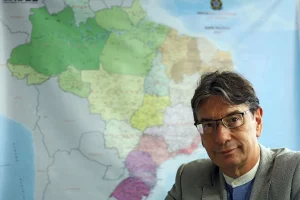Homem de terno e óculos em frente a mapa do Brasil, representando dados do IBGE e informações geográficas.