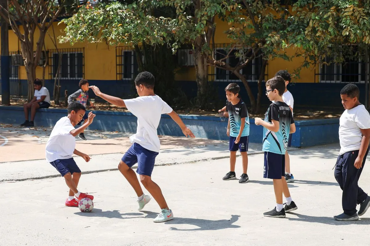 Crianças jogam futebol no pátio de escola pública durante o recreio, em área externa arborizada.