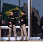 Policiais armados realizam segurança em frente a prédio público com bandeira do Brasil refletida no vidro.