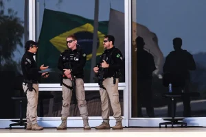 Policiais armados realizam segurança em frente a prédio público com bandeira do Brasil refletida no vidro.