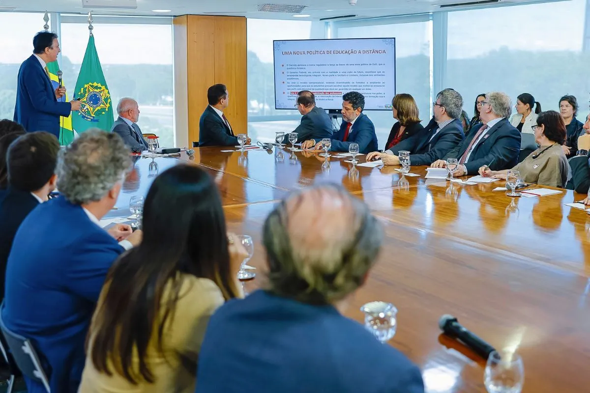 Autoridades do governo federal participam de reunião ministerial sobre política de educação a distância, com apresentação em tela e bandeira do Brasil ao fundo.