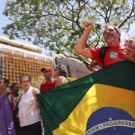 Manifestantes seguram bandeira do Brasil durante protesto político ao ar livre, com pessoas fazendo gestos e palavras de ordem.