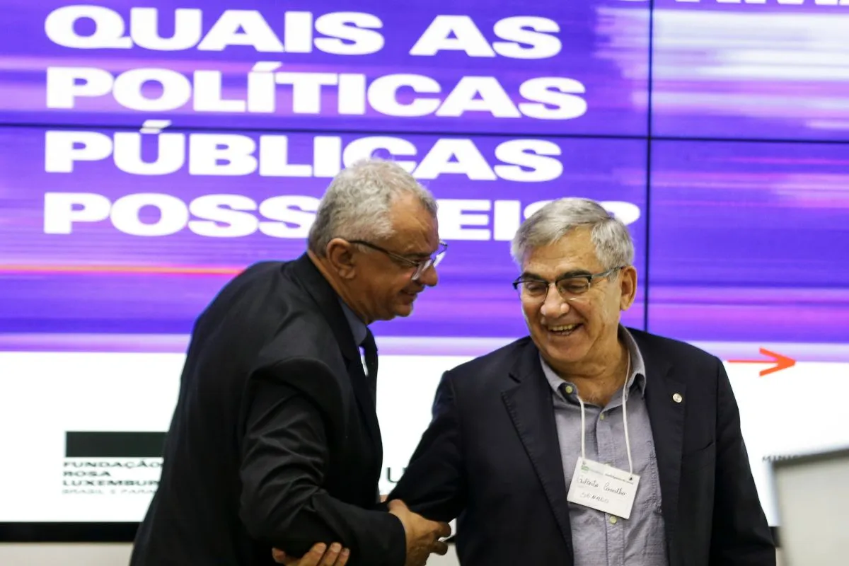 Dois especialistas apertam as mãos durante evento sobre políticas públicas com painel ao fundo.