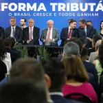 Autoridades políticas participam de evento institucional sobre reforma tributária, com público e painel oficial ao fundo.