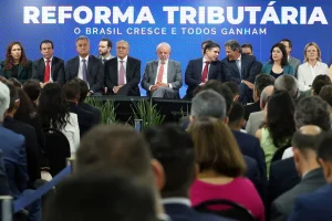 Autoridades políticas participam de evento institucional sobre reforma tributária, com público e painel oficial ao fundo.