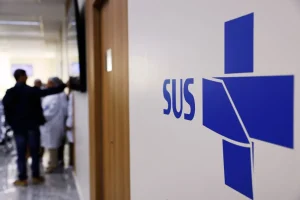 Logotipo do SUS em parede de hospital público com profissionais de saúde ao fundo.