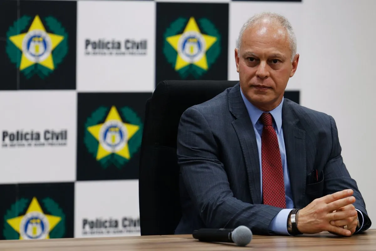 Delegado da Polícia Civil sentado em mesa durante coletiva de imprensa sobre investigação criminal.