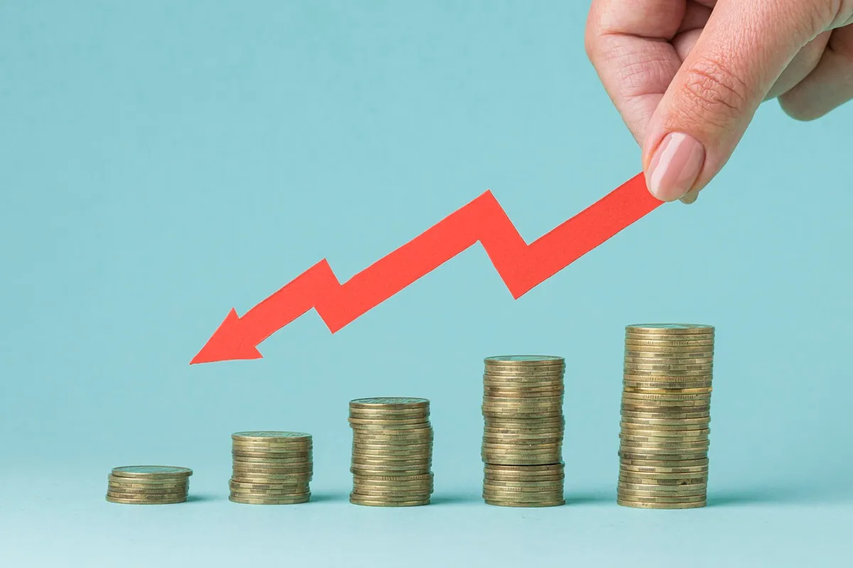 Mão segurando seta vermelha apontando para baixo sobre pilhas crescentes de moedas, representando queda na taxa de juros ou redução de índice econômico.