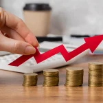 Mão segurando seta vermelha apontando para cima sobre pilhas crescentes de moedas, simbolizando aumento da taxa de juros e crescimento econômico.
