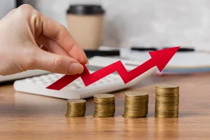 Mão segurando seta vermelha apontando para cima sobre pilhas crescentes de moedas, simbolizando aumento da taxa de juros e crescimento econômico.