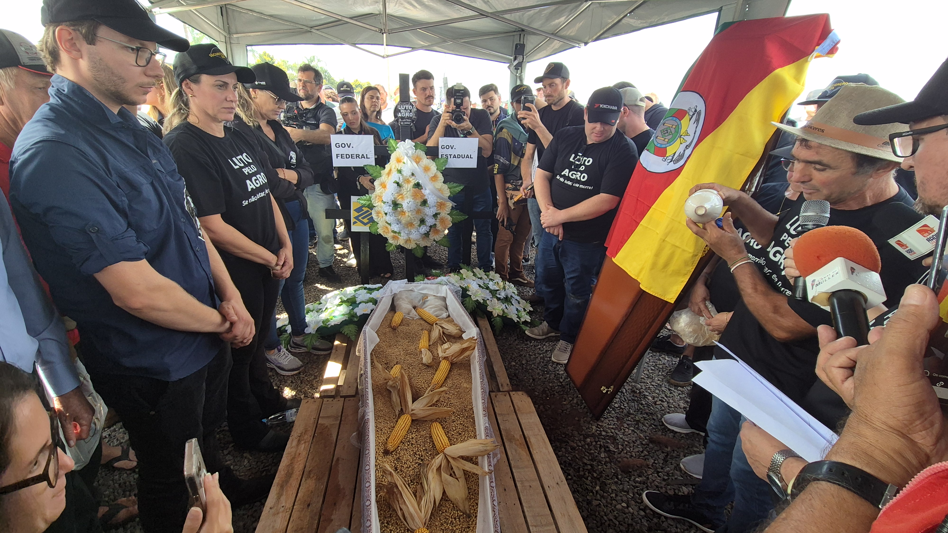Produtores rurais despejam um pouco de soja, milho, trigo e uma garrafa de leite em um caixão, um ato que simboliza a 'morte' do agronegócio, junto de placas com os nomes do governo estadual, federal, bancos e cooperativas de crédito. O protedto ocorreu na abertura da 26ª Expodireto Cotrijal, uma das maiores feiras de agronegócio da América Latina | Foto: Tauany Cattan/Revista Oeste