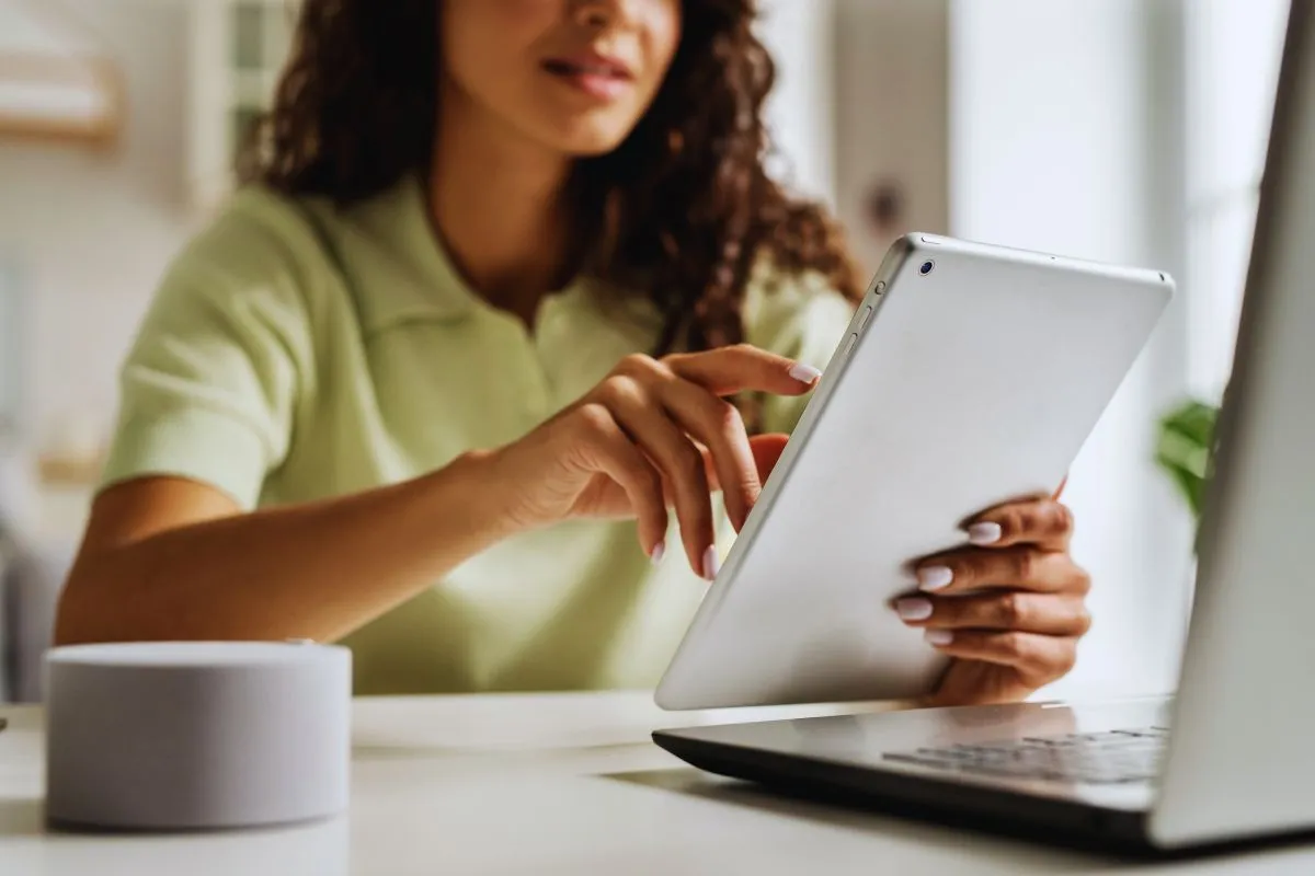 Mulher usando tablet em mesa de trabalho ao lado de notebook e xícara de café.