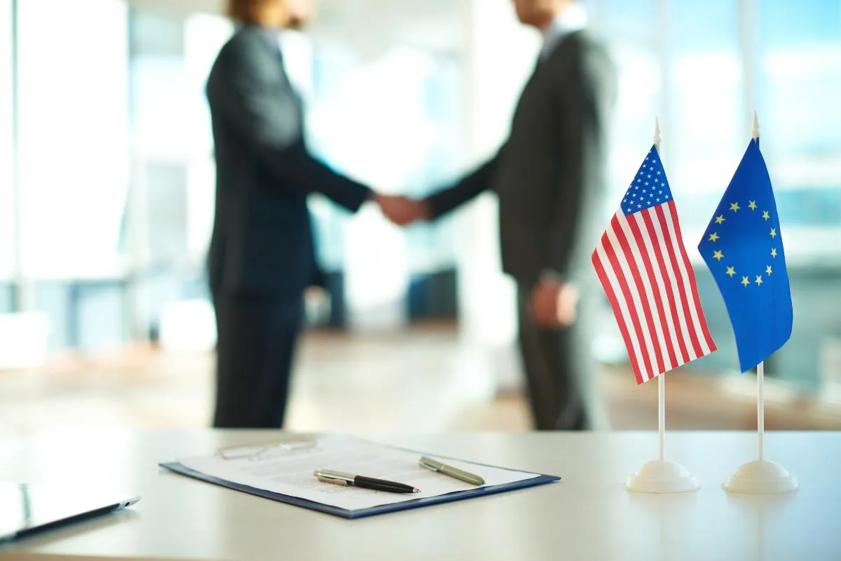 Bandeiras dos Estados Unidos e da União Europeia sobre mesa com empresários apertando as mãos ao fundo