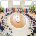 Líderes de diversos países reunidos em mesa oval com bandeiras internacionais ao fundo