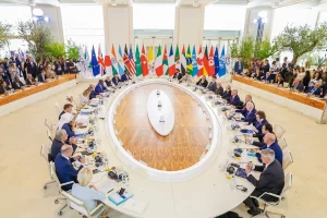 Líderes de diversos países reunidos em mesa oval com bandeiras internacionais ao fundo