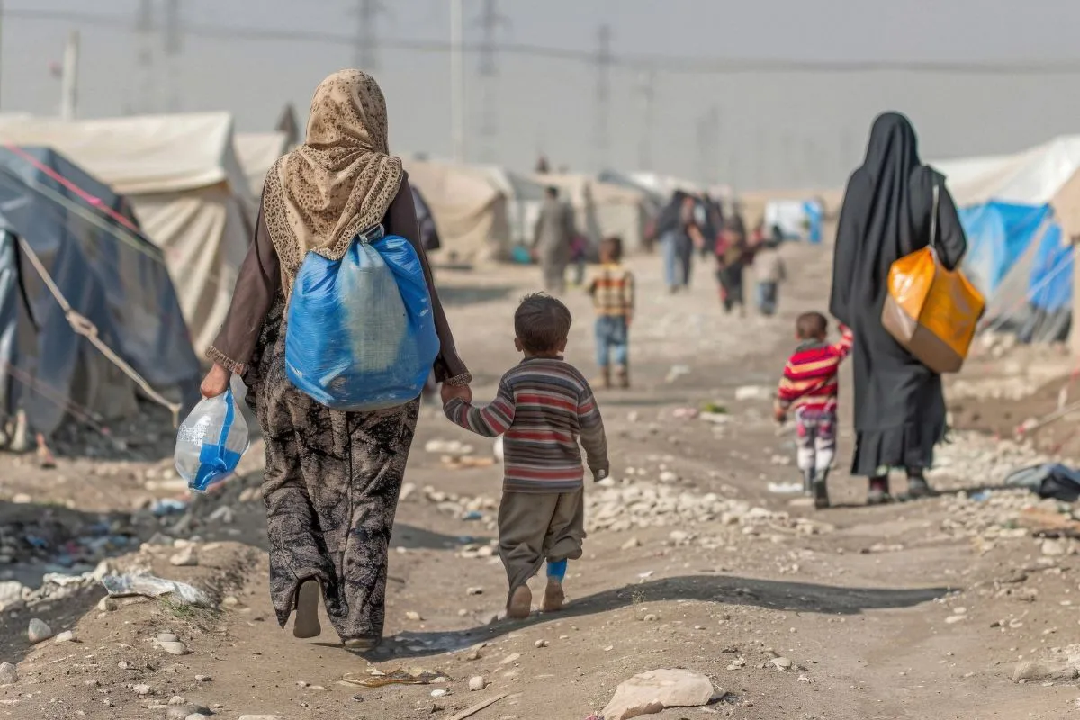 Mulher e criança caminhando em campo de refugiados com tendas ao fundo em cenário de crise humanitária