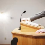 Homem discursando em púlpito com microfone e livro aberto em ambiente institucional
