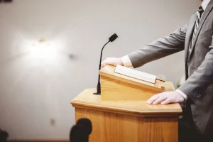 Homem discursando em púlpito com microfone e livro aberto em ambiente institucional