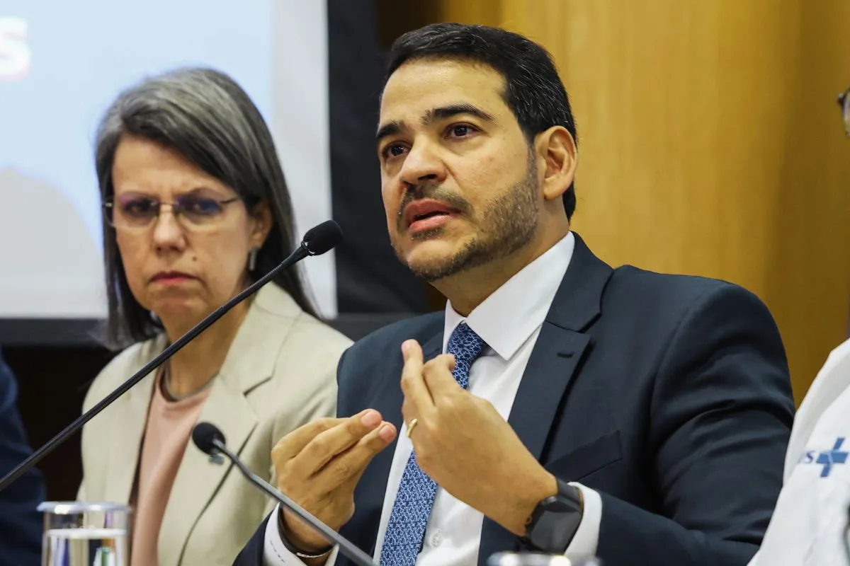 Homem de terno falando em coletiva de imprensa com microfone, ao lado de mulher em evento oficial sobre economia e gestão pública.